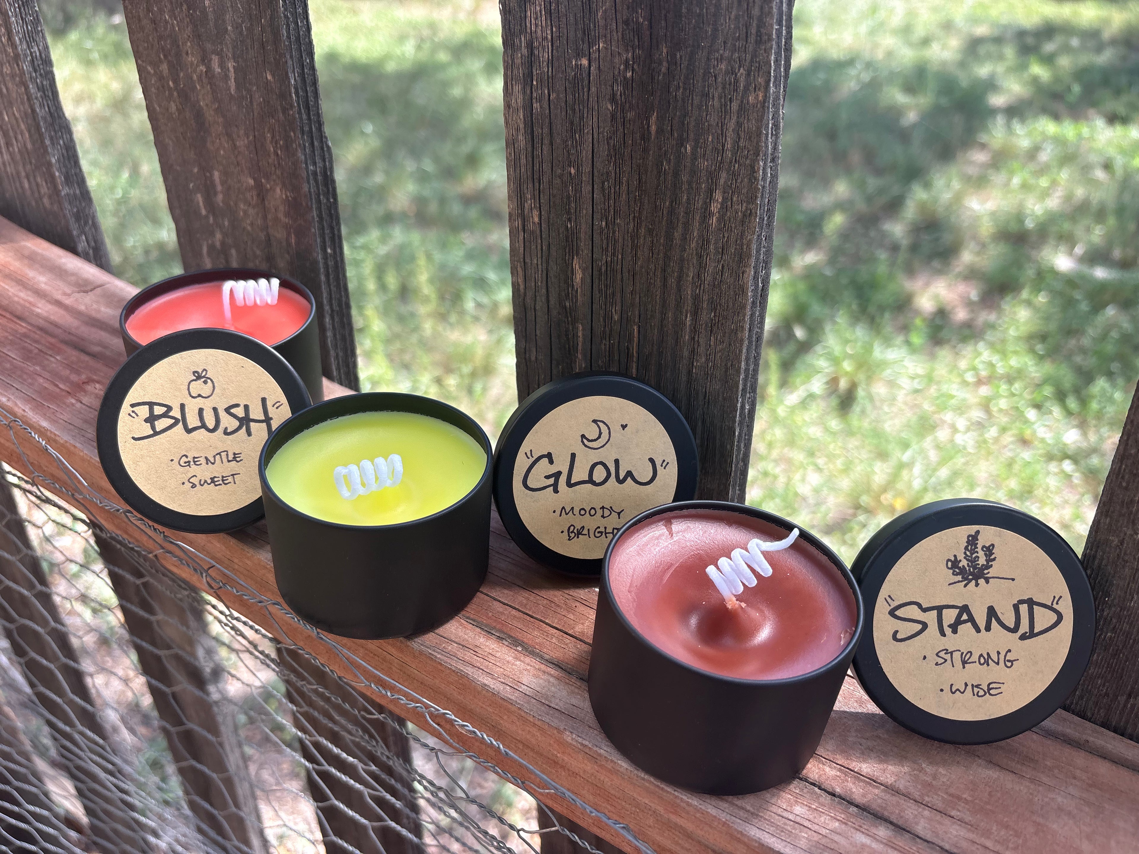 Four scented candles in round black metal tins with beige labels, arranged on a wooden deck railing outdoors. The labels read Blush, Glow, Stand, and Mostly Bright, with wicks visible in the open tins.